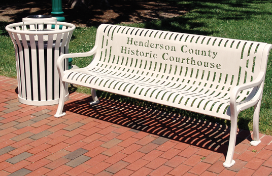 White bench and receptacle for Henderson County Historic Courthouse on a brick sidewalk in front of grass.