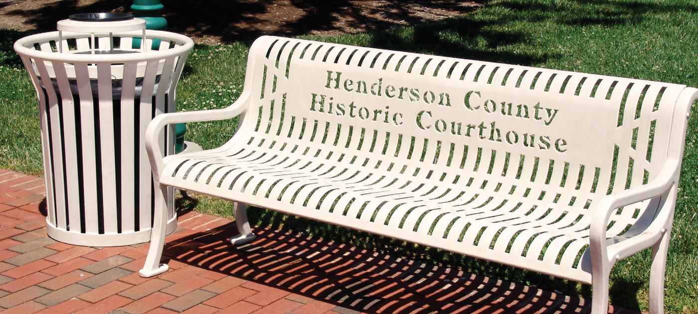 White bench and receptacle for Henderson County Historic Courthouse on a brick sidewalk in front of grass.